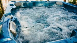 A blue hot tub is shown with bubbling water, surrounded by wooden decking and a lush natural backdrop. The hot tub is designed to accommodate multiple people, providing a space for relaxation and enjoyment.
