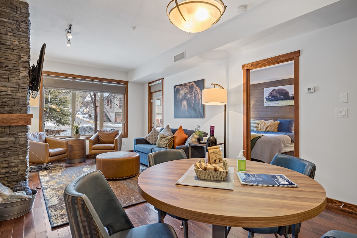 A warm living area features two comfortable seating arrangements, a textured area rug, and a cozy dining table with stylish chairs. Natural light fills the space through large windows, offering glimpses of the outdoors. Two doors lead to separate bedrooms, enhancing the layout's privacy.