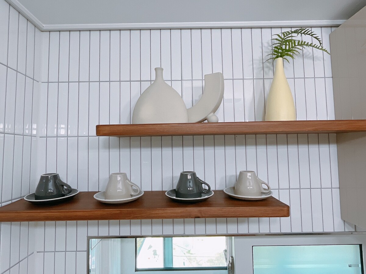 Open shelves display an assortment of ceramic cups and saucers in neutral tones. A decorative arrangement of modern vases and a green plant add to the design above. The backdrop features white tiles with a subtle vertical pattern.