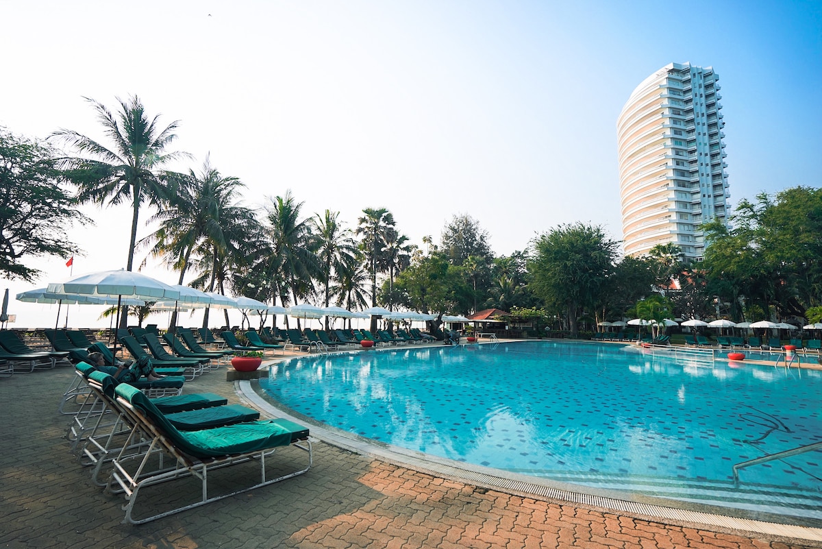 An expansive pool area is surrounded by lush palm trees and lounge chairs. A tall building is visible in the background, reflecting the tranquil setting. Sun umbrellas provide shaded spots for relaxation beside the clear water of the pool.