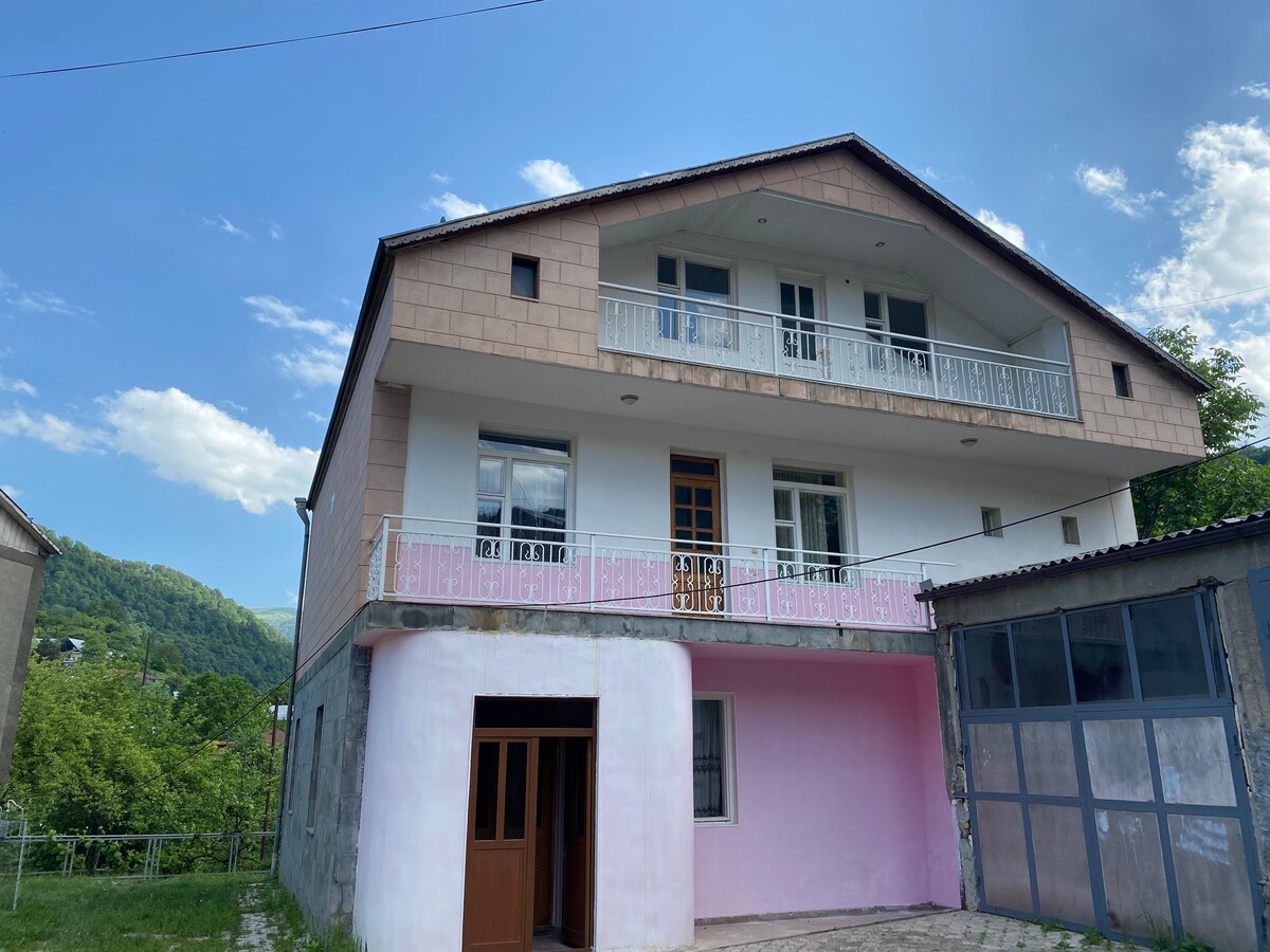 The two-story house is set against a backdrop of green hills, featuring a light-colored façade and a spacious balcony. The entrance is marked by double doors, and multiple windows allow natural light to illuminate the interior.