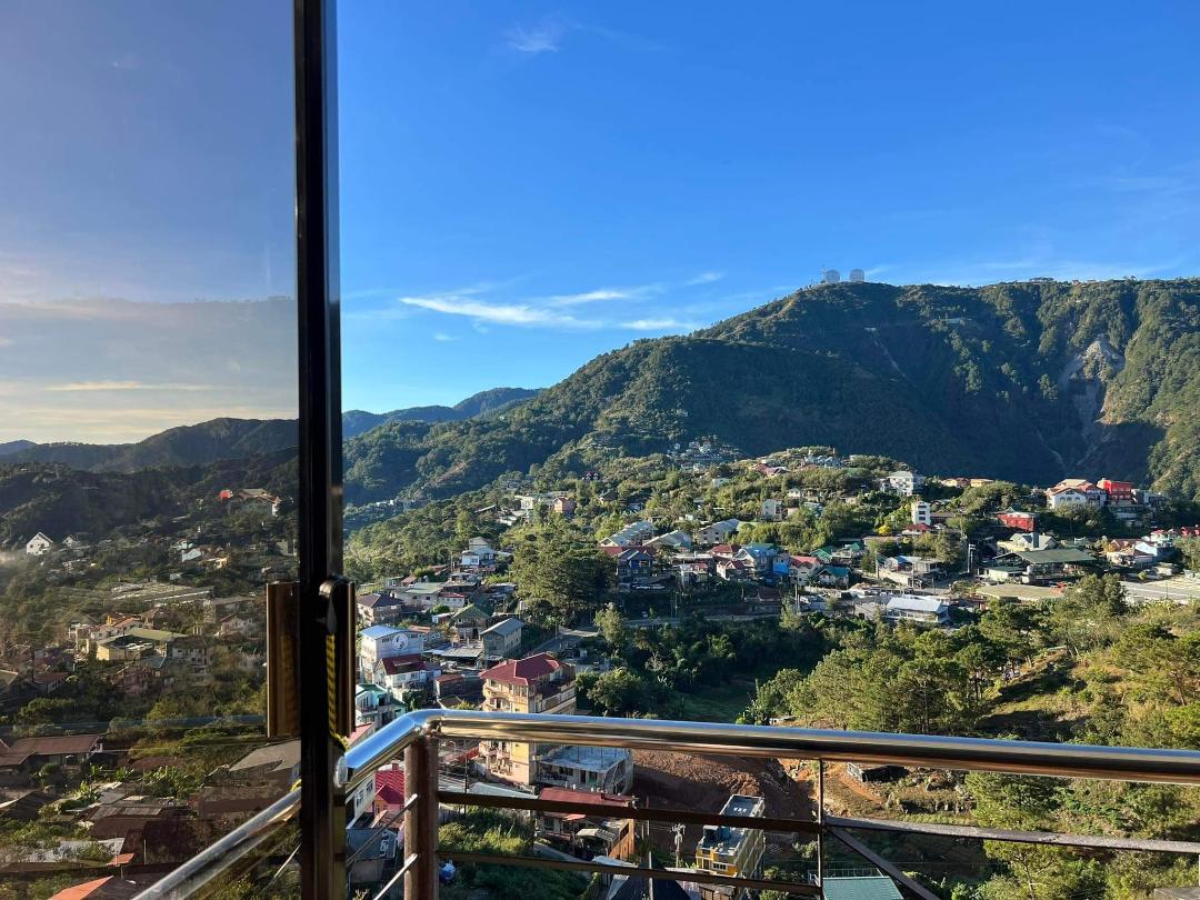 A corner balcony offers expansive views of lush mountains and colorful houses below, framed by clear skies. The landscape showcases varied greenery, with visible roofs of nearby homes nestled within the mountainside.