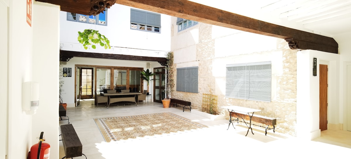 A spacious courtyard is depicted with a stone floor and large windows allowing natural light to fill the area. Benches are positioned along the walls, and a defined seating area is featured in the center, surrounded by greenery and stone walls.
