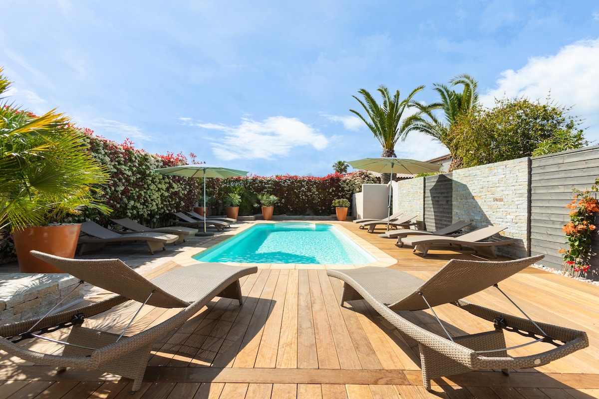 A secure, heated shared pool area is surrounded by lush greenery and colorful flowering plants. Multiple sun loungers are positioned around the wooden deck, inviting relaxation under the sun, while umbrellas provide shaded spots for comfort.