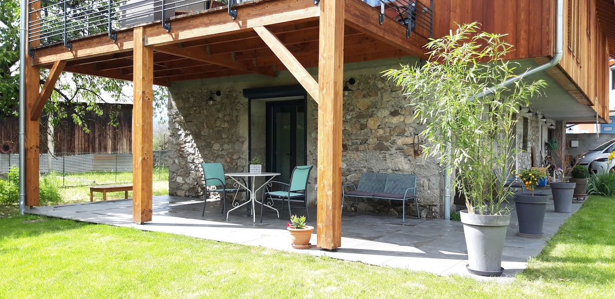 A terrace is showcased beneath a wooden structure, featuring outdoor seating with a small table and two chairs. Potted plants and a well-maintained lawn complete the inviting space, with stone walls visible in the background.