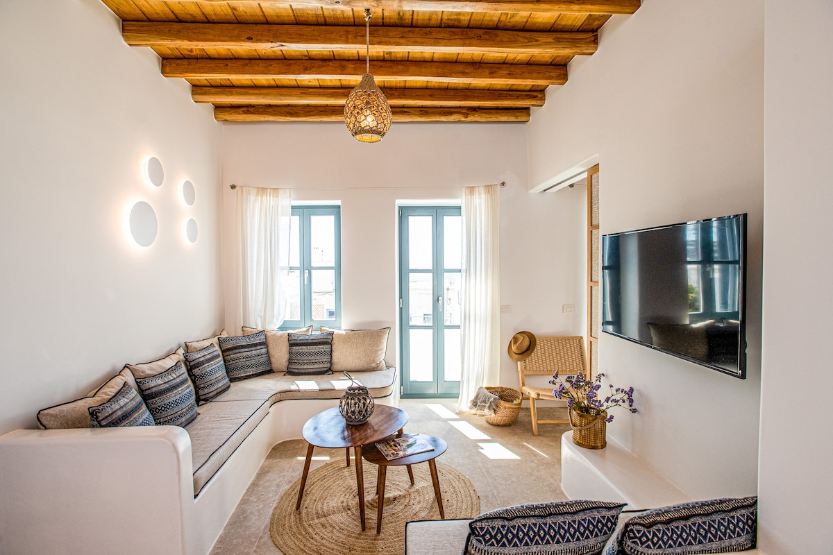 A spacious living area features a large L-shaped sofa adorned with cushions, complemented by a round wooden coffee table. Natural light enters through two windows framed with sheer curtains, and a flat-screen TV is mounted on the wall. Wooden beams add warmth to the ceiling.