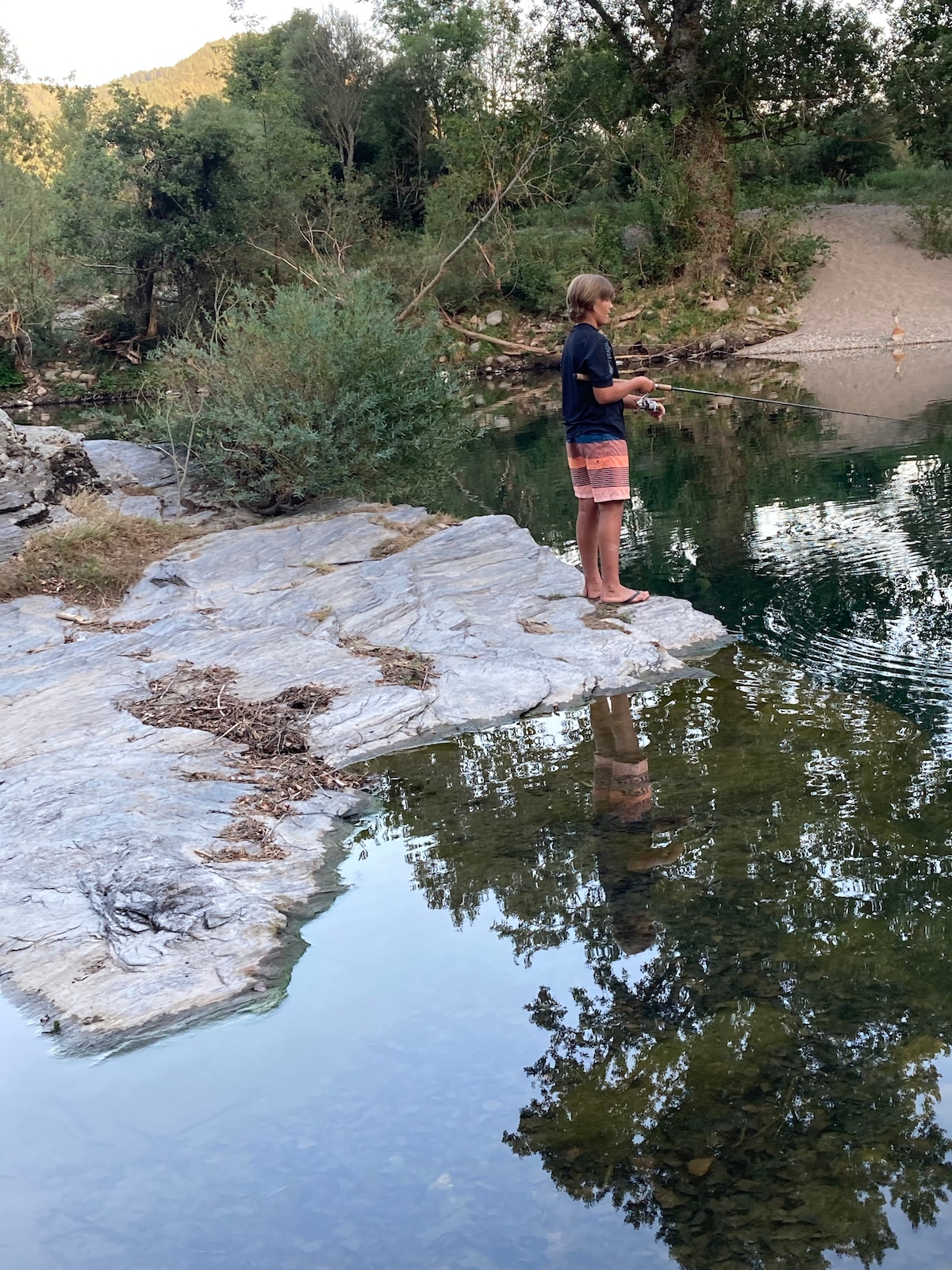A young individual stands barefoot on a large rock, casting a fishing line into calm waters. Surrounding foliage and trees reflect, creating a serene atmosphere. The background features a sandy area visible beyond the trees, suggesting a peaceful natural setting.