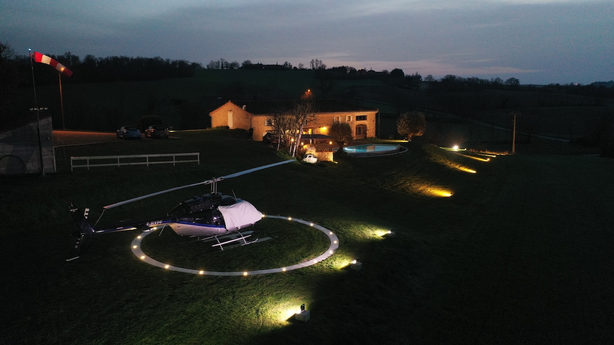An aerial view captures a helicopter positioned on a well-lit landing pad, surrounded by landscaped grounds. A luxurious house with soft exterior lighting stands alongside a serene pool, framed by lush greenery and rolling hills under a twilight sky.