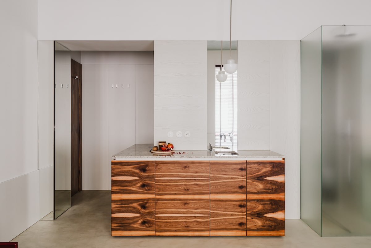 The kitchen features a modern wooden island with a smooth countertop and minimalist design. Glass panels extend along the walls, allowing natural light to brighten the space. The open layout emphasizes a clean and functional aesthetic, highlighting the contemporary finishes and fixtures.