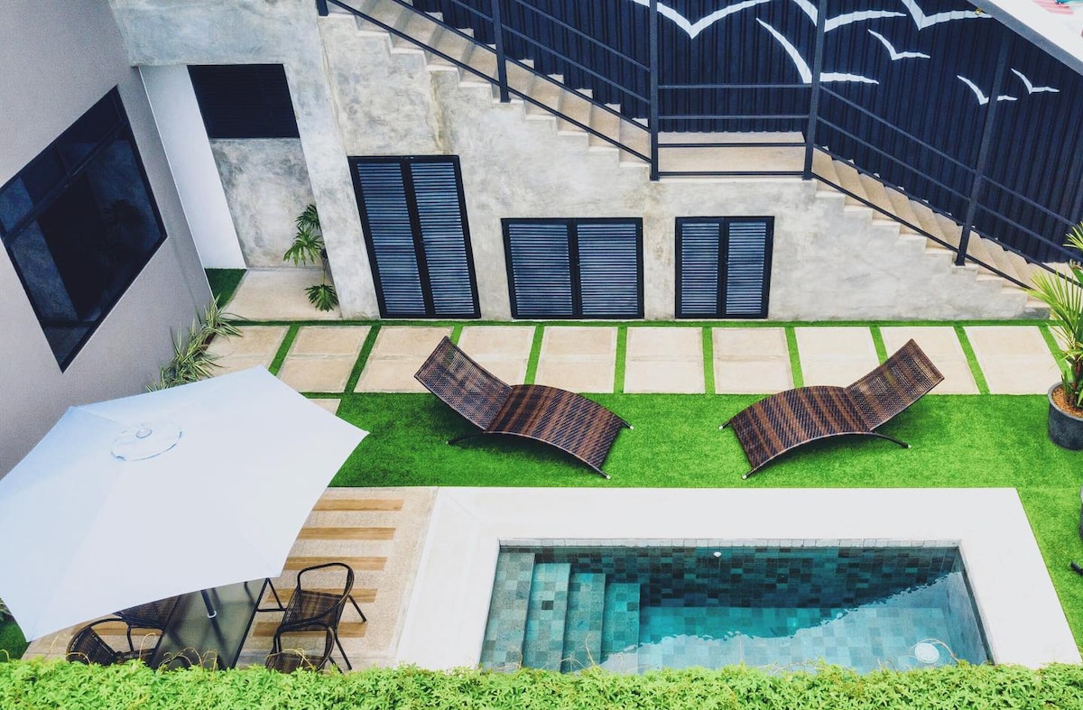 An aerial view of the outdoor area showcases a natural stone pool surrounded by lush greenery. Two lounge chairs are positioned on the grass, and a patio umbrella provides shade over a small seating area with chairs and a table.