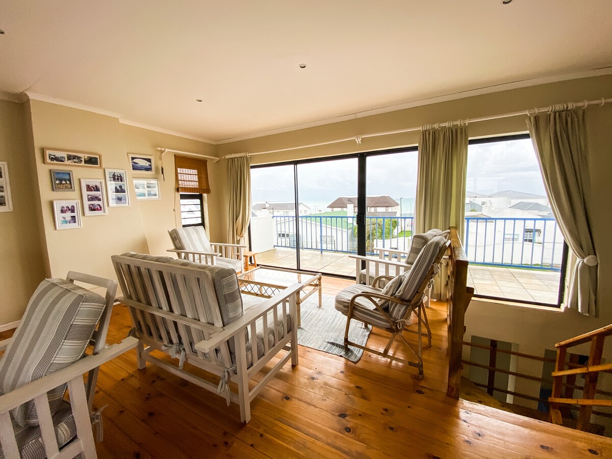 A spacious family room features multiple seating options, including two lounge chairs and a coffee table. Large windows provide natural light and showcase views of the balcony. Light-colored curtains frame the windows, while wooden flooring adds warmth to the space.