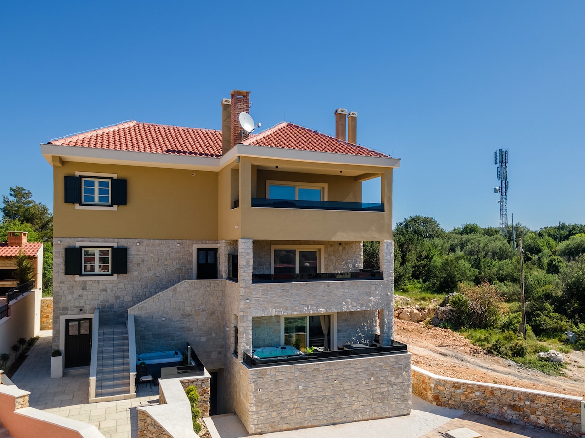 The modern duplex structure features a combination of stone and contemporary elements. Balconies extend out from the upper level, offering views of the surrounding greenery. Large windows invite natural light into the living spaces, while a jacuzzi is visible on the lower terrace.