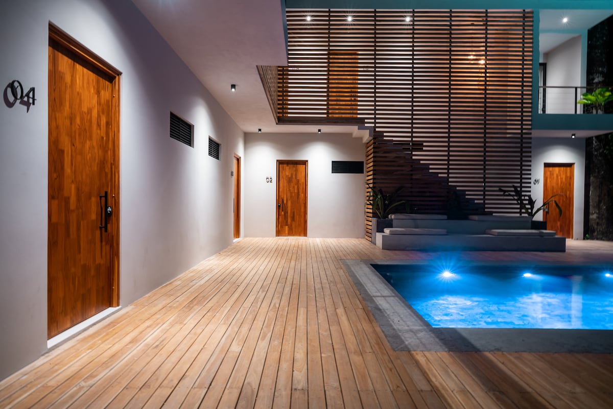 A well-lit pathway leads to several wooden doors, each marked with numbers. The contemporary design includes a softly glowing pool at one side, surrounded by smooth stone edges, and a wooden deck extending throughout the area. A staircase is positioned in the background, enhancing the open layout.
