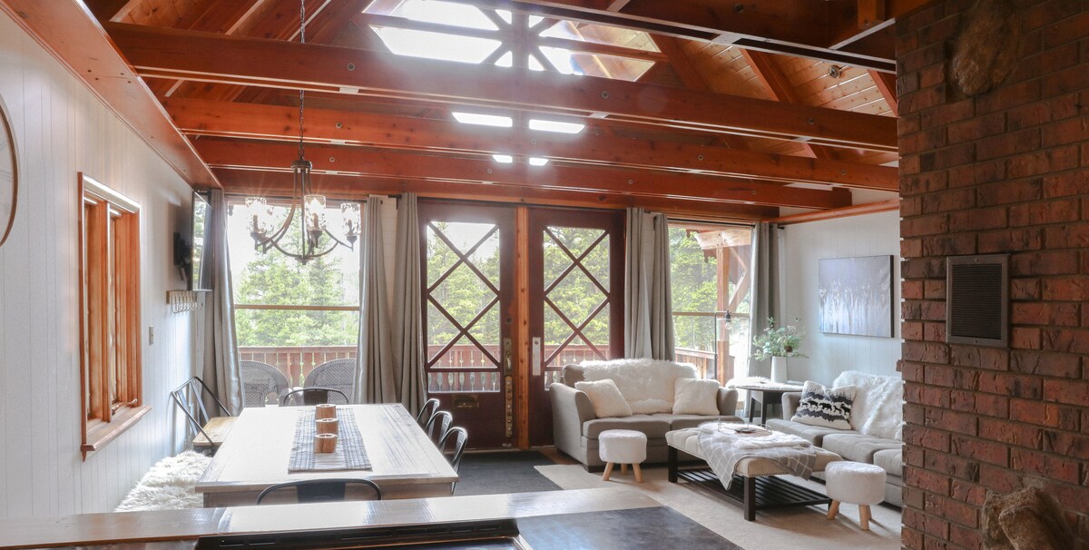 An open living space showcases wooden beams above, with large windows allowing natural light to fill the room. A dining table is positioned centrally, surrounded by chairs. A comfortable seating area includes plush sofas and a cozy rug, with a view of the outdoors visible through the glass doors.
