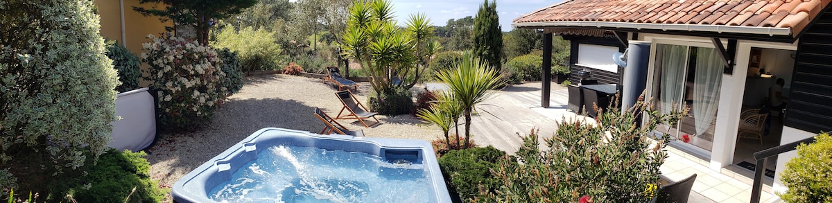 An inviting outdoor space is featured, showcasing a hot tub surrounded by lush greenery and landscaped garden elements. Comfortable lounge chairs are positioned nearby, and a terrace with dining options is partially visible. The peaceful setting offers a serene atmosphere with scenic views.