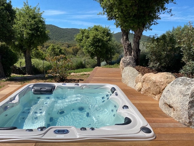 A jacuzzi with multiple seating areas is positioned on a wooden deck. Lush greenery and rolling hills are visible in the background, enhanced by a clear blue sky. Natural stone elements complement the tranquil outdoor setting.