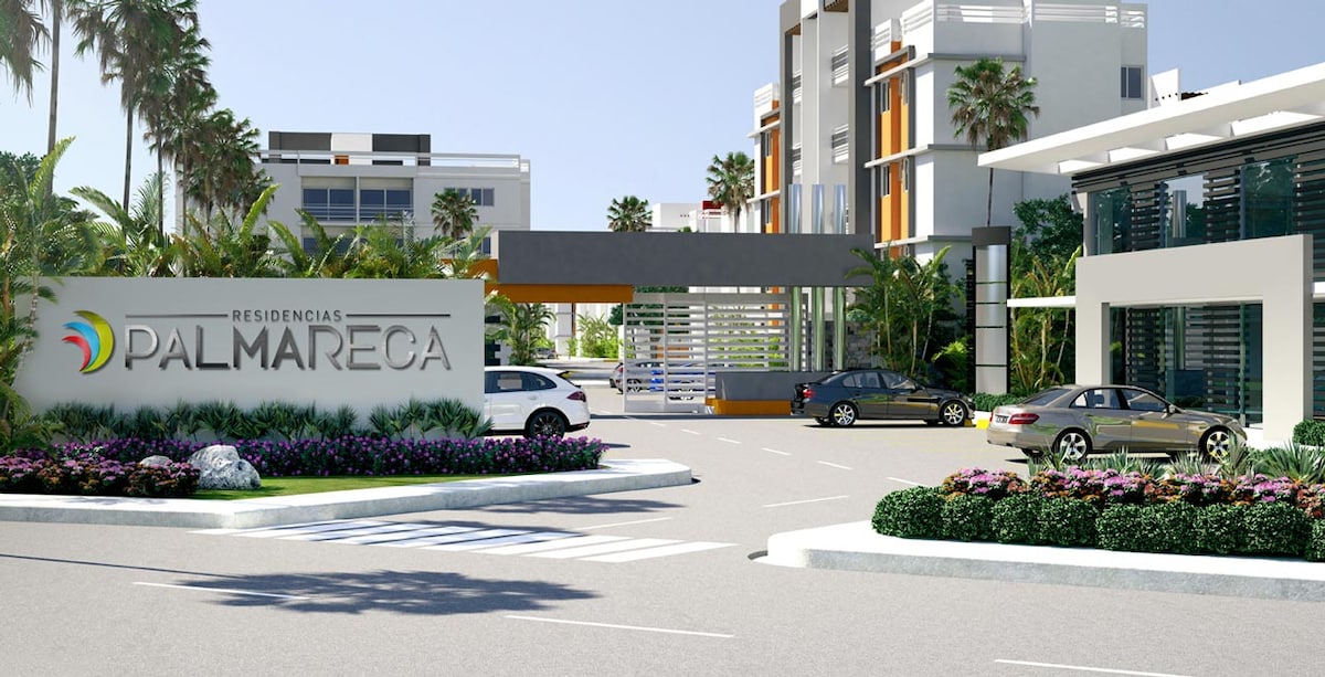 The entrance of Residencias Palmareca is depicted, featuring a stylish sign amidst landscaped greenery. Palm trees line the driveway, while modern buildings are visible in the background. Several parked cars can be seen, indicating a welcoming and accessible environment.