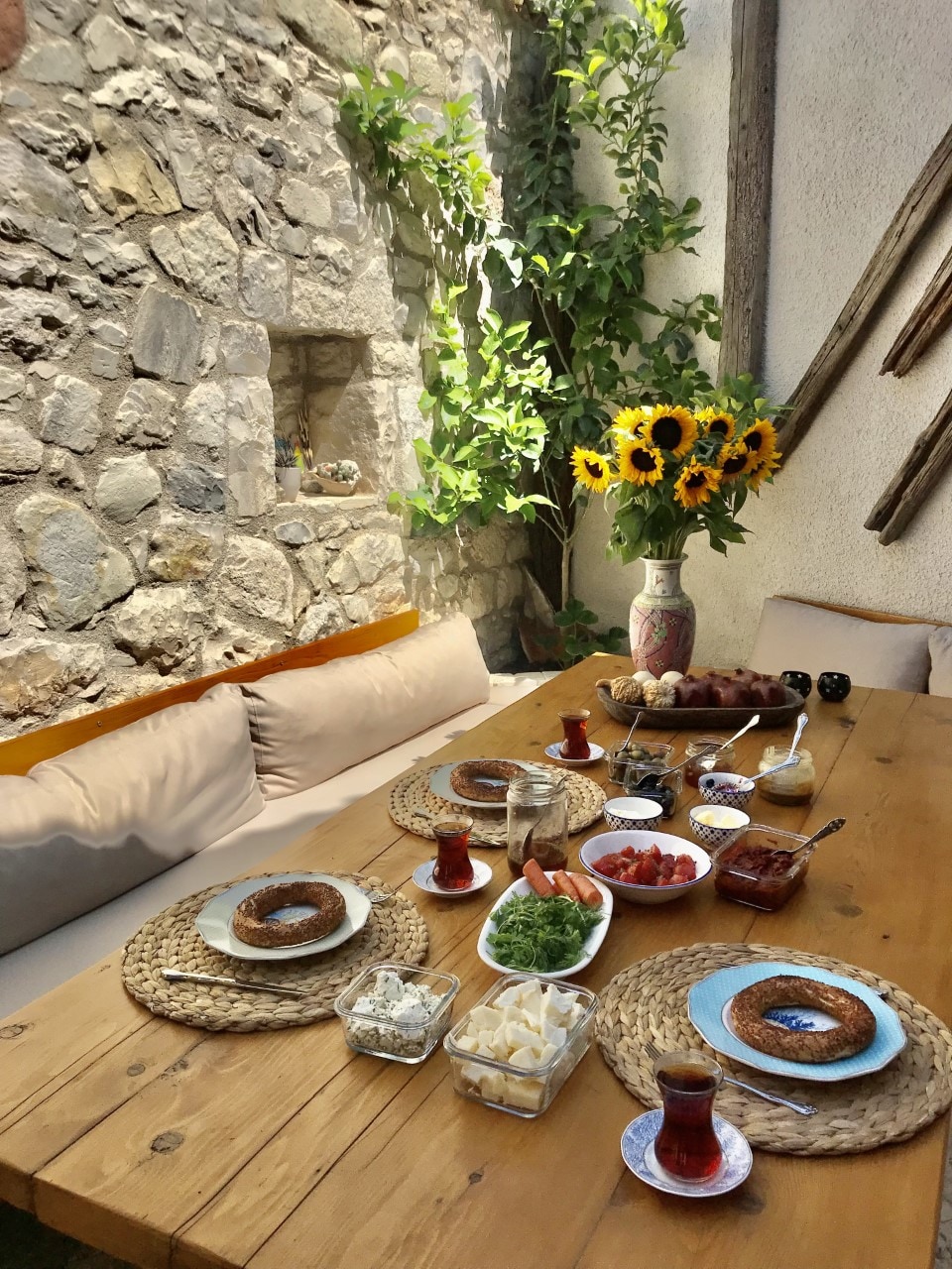 A wooden dining table is set for a meal in a sunlit courtyard, surrounded by stone walls. Plates filled with fresh produce, cheese, and bread are arranged alongside glasses of tea. Bright sunflowers and greenery offer a natural touch, creating a serene dining atmosphere.