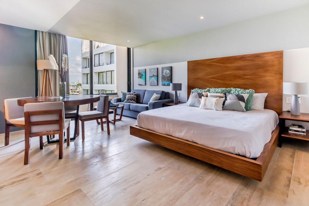 A spacious studio features a king-size bed with a wooden base, complemented by decorative pillows. A dining area with four chairs and a round table adjoins a comfortable sofa. Natural light illuminates the room through large windows, enhancing the neutral color palette.