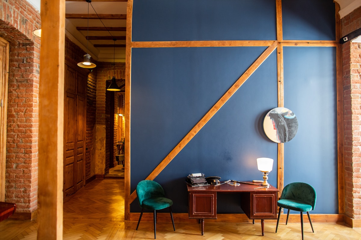 A section of the interior showcases a deep blue accent wall enhanced by wooden beams. Two green chairs are positioned near a stylish console table, which holds a vintage lamp and a round mirror, reflecting the inviting space.