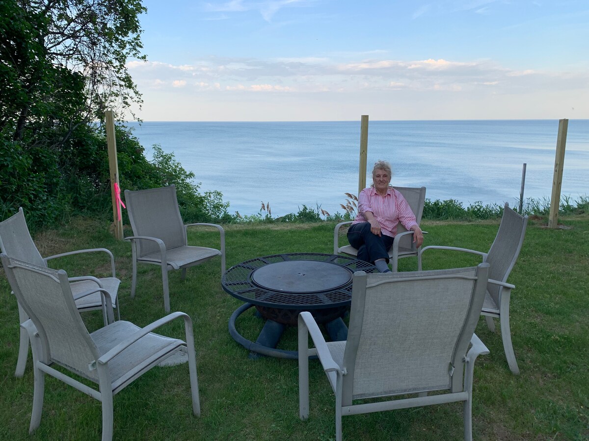 A cozy fire pit area is set in a grassy space, surrounded by six comfortable chairs. The scene offers a clear view of Lake Huron, with calm waters stretching to the horizon under a light blue sky.