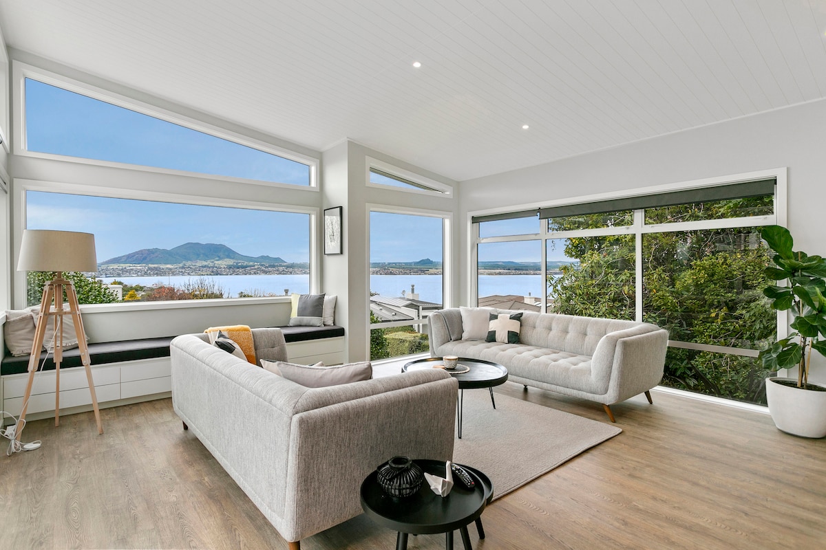 A modern living space is defined by large windows that frame expansive lake views, allowing for abundant natural light. A cozy seating arrangement includes a light gray sectional sofa and a two-tone coffee table, while a stylish floor lamp complements the overall design.