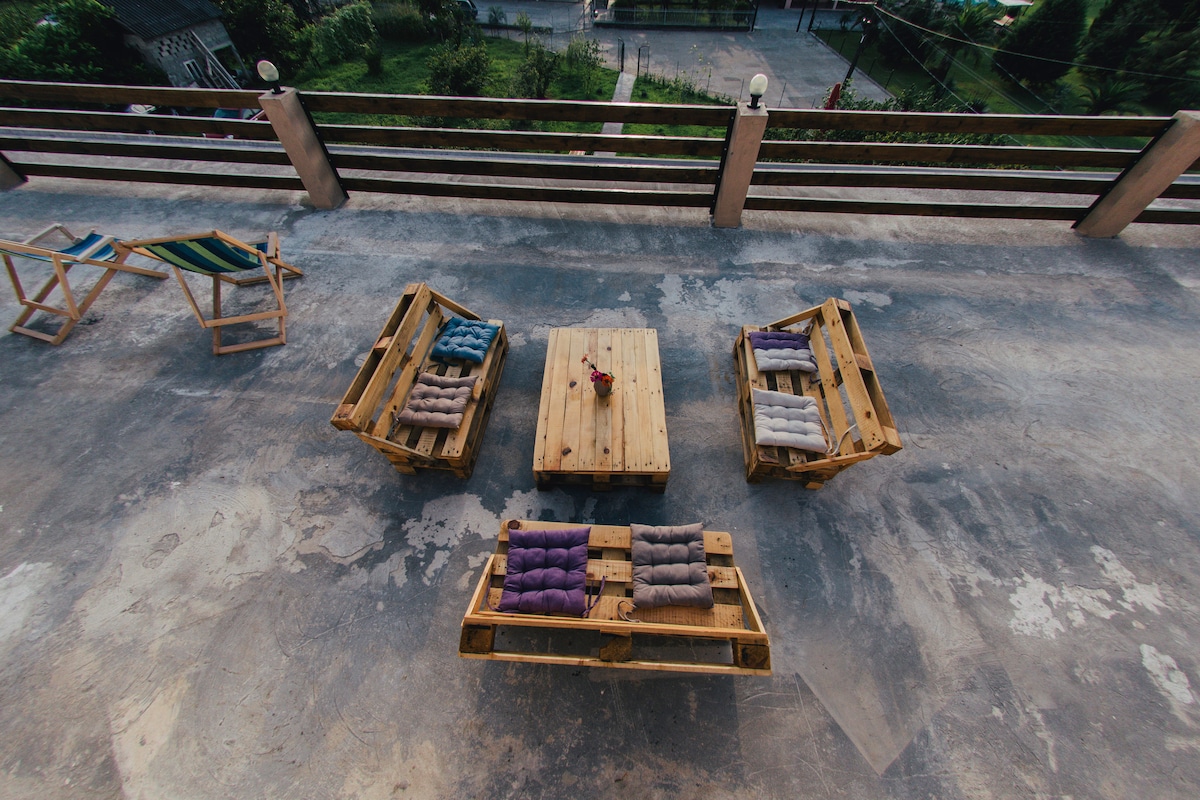 An outdoor seating area features wooden pallet furniture arranged in a circle. Colorful cushions adorn the seats, alongside a wooden table in the center. A view of the surrounding greenery and a driveway is visible beyond the railing, enhancing the inviting atmosphere.