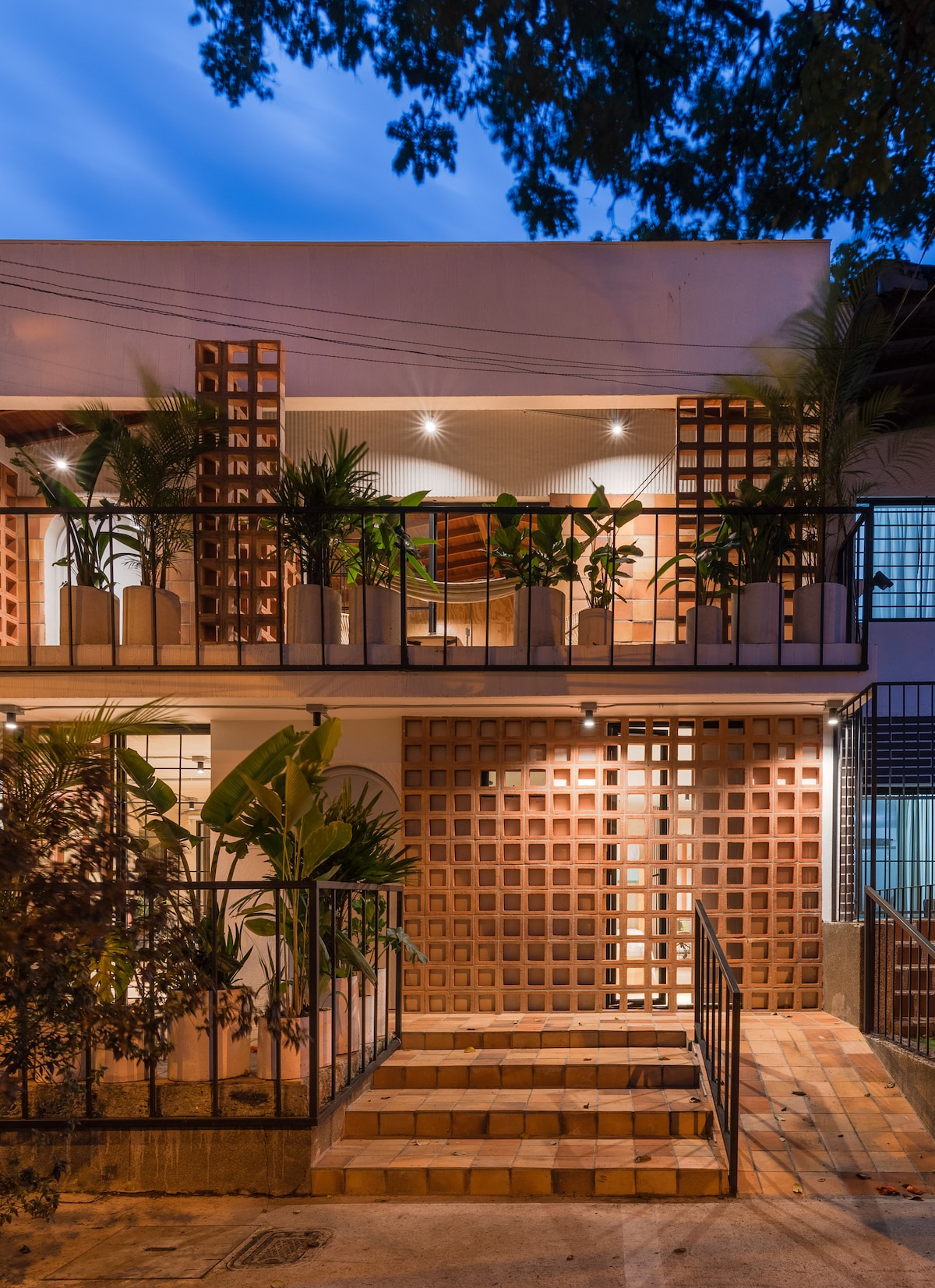 The exterior of the apartment features a modern design with a combination of decorative brickwork and abundant greenery. Potted plants line the steps leading to the entrance, illuminated by warm lighting that enhances the welcoming atmosphere. The balcony offers a glimpse of the interior through large windows.