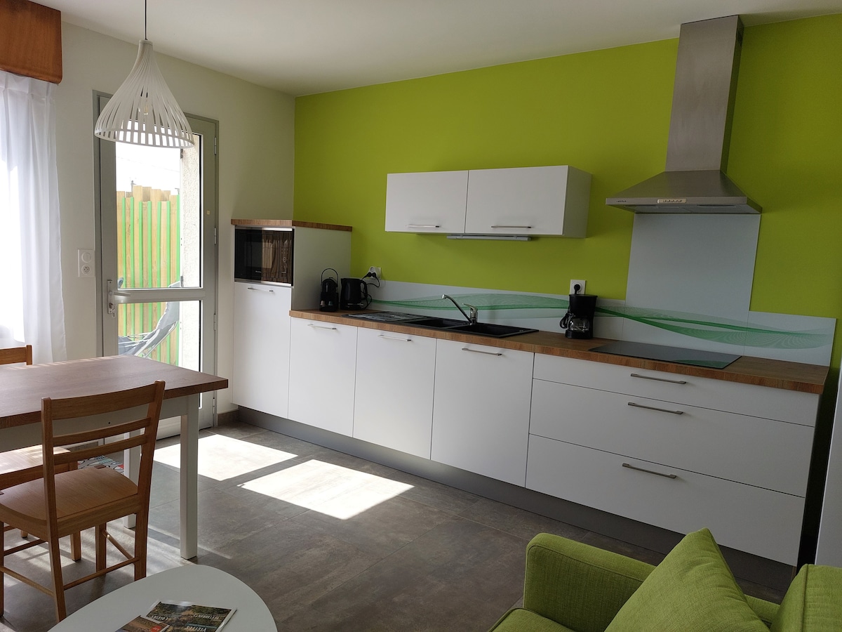 A bright kitchen area features a vibrant green accent wall complemented by sleek white cabinetry. A countertop with cutting board and kitchen appliances, including a microwave and coffee maker, is visible. A wooden dining table with four chairs, alongside a green sofa, creates a functional living space.
