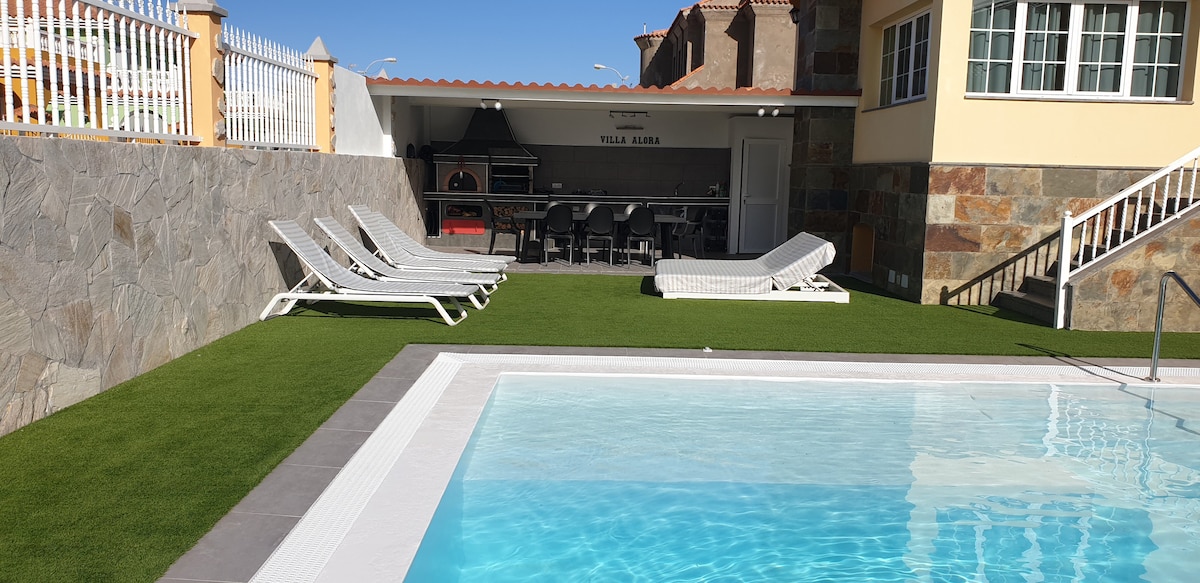 A heated swimming pool is surrounded by well-maintained artificial grass. Sun loungers are positioned nearby for relaxation. The outdoor space is paired with a covered area featuring a barbecue grill and seating, providing an inviting space for gatherings.