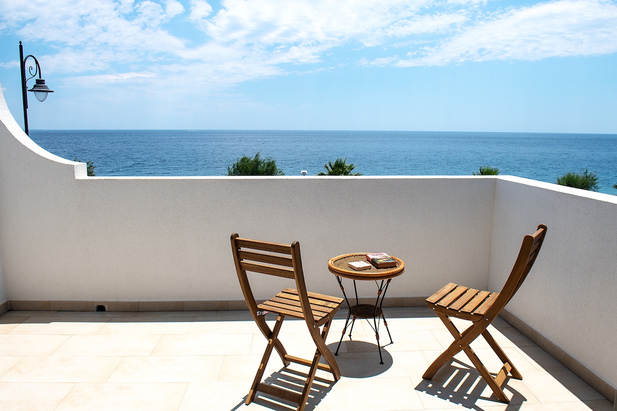A sunlit terrace presents stunning sea views, furnished with two wooden folding chairs and a small round table. The light-colored tiles create a spacious feel, while the open sky and water enhance the serene atmosphere.