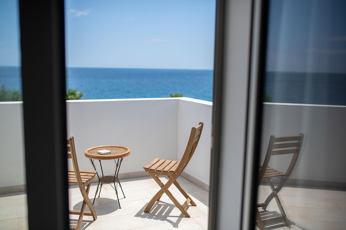 A balcony is presented with a clear view of the sea. Two wooden chairs and a small round table are arranged on the tiled surface, creating a serene space for relaxation. The ocean glimmers under bright sunlight in the background.