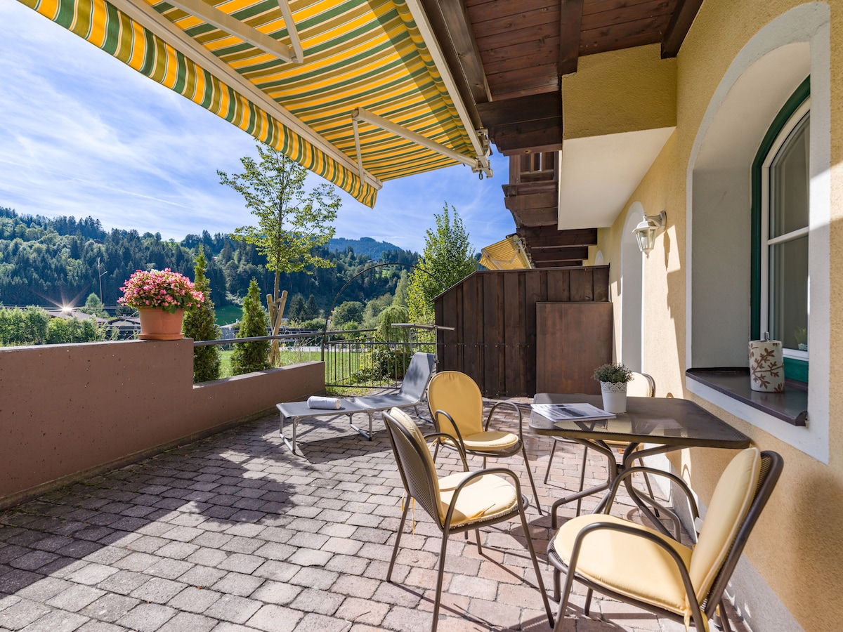 An outdoor terrace features yellow-striped awnings providing shade over a small round table and two chairs. A sun lounger is positioned nearby, and potted flowers add a touch of color. The view showcases rolling green hills and a clear blue sky.
