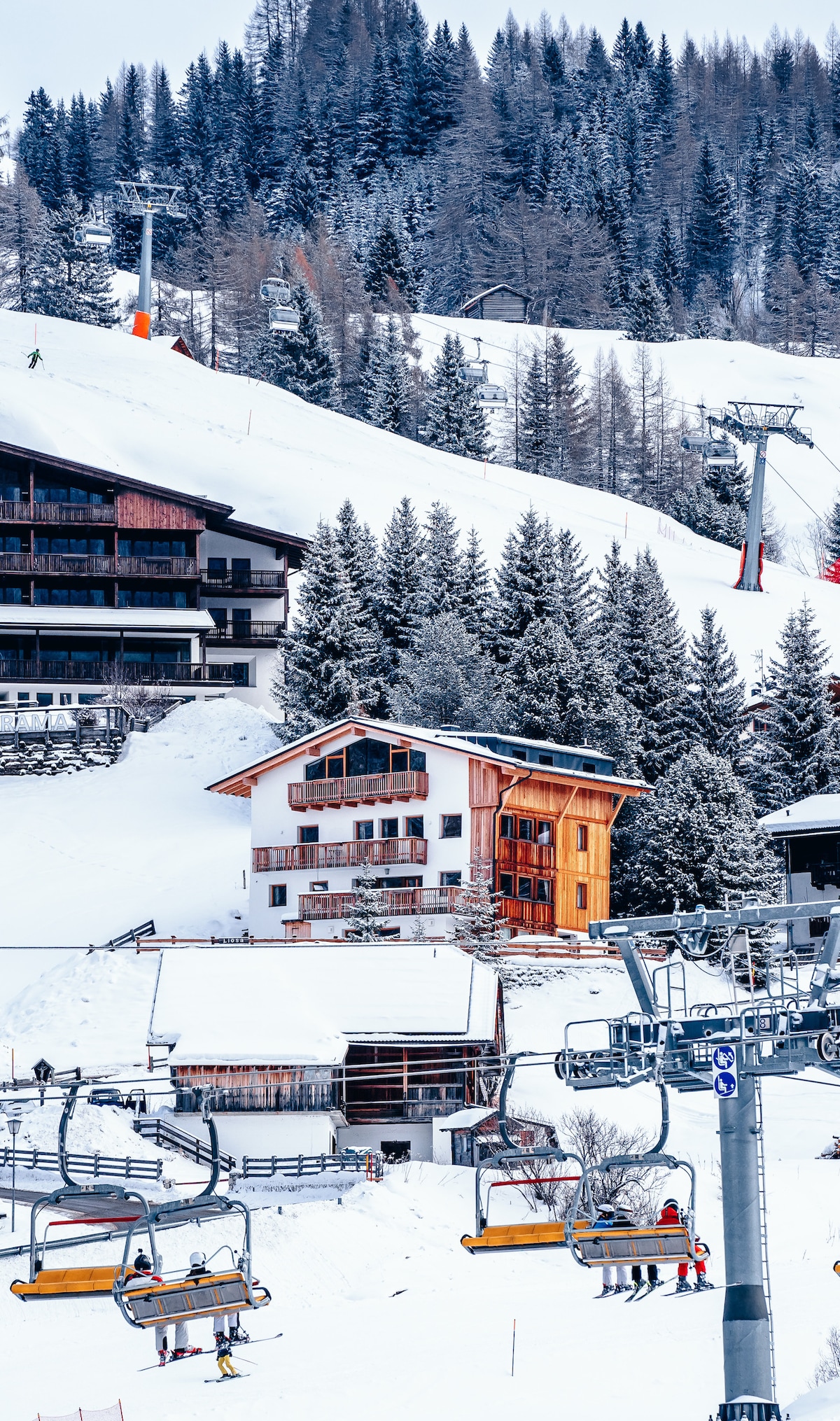 A wooden chalet stands prominently amidst snowy slopes, surrounded by tall evergreen trees. Ski lifts are visible nearby, transporting guests to the snow-covered mountains above. The scene captures the essence of a winter alpine landscape, with soft layers of white snow blanketing the ground.