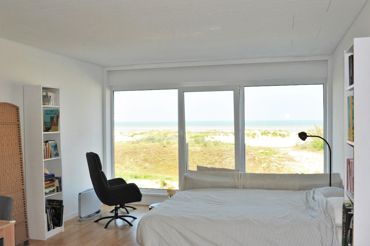 A spacious studio features large windows offering direct sea views. A comfortable double bed is present, alongside a cozy armchair. A bookshelf is positioned against one wall, contributing to the airy atmosphere. Natural light brightens the room, creating a serene coastal ambiance.