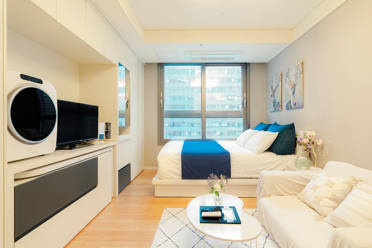 A well-lit room features a spacious bed with a blue accent blanket, complemented by decorative pillows. Natural light enters through large windows, while a comfortable seating area is arranged with a light-colored couch and a small coffee table.