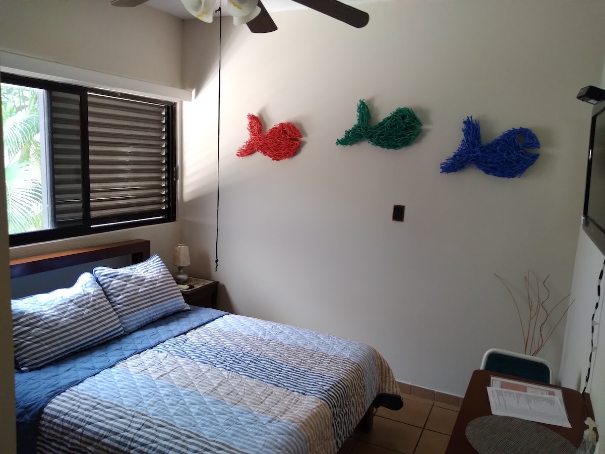 A cozy sleeping area is presented with a spacious bed dressed in a quilted coverlet. Above the bed, three decorative fish in vibrant red and blue tones adorn the wall. A ceiling fan hangs overhead, and a small desk with a chair is situated nearby.