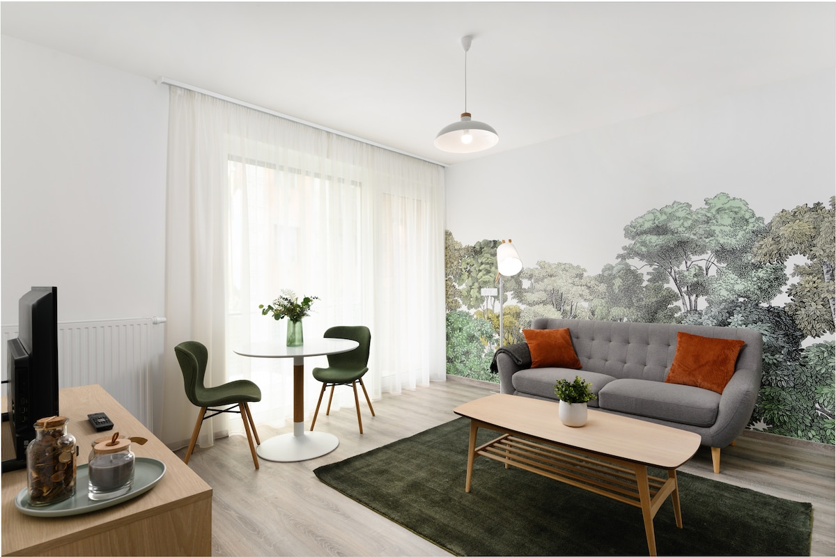 A modern living area features a gray sofa with orange cushions, paired with two green chairs around a circular table. A coffee table sits centrally, and a plush green rug covers the floor. Natural light enters through sheer curtains, illuminating a mural of greenery on the wall.