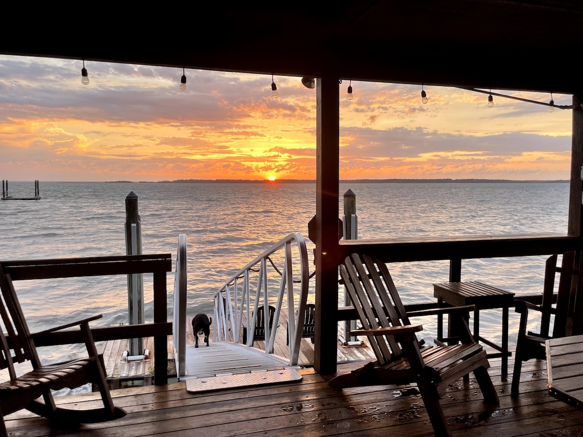 A view of the sunset over the water is captured from a covered deck. Wooden seating, including Adirondack chairs, is positioned for relaxation. A private dock leads to the water, and soft lighting from hanging bulbs adds to the serene setting.