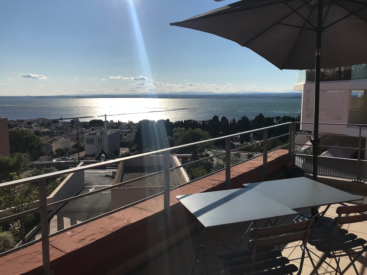 An expansive terrace is shown with a view overlooking the water, illuminated by sunlight. A table and several chairs are positioned for outdoor dining. An umbrella provides shade, enhancing comfort while enjoying the serene landscape.