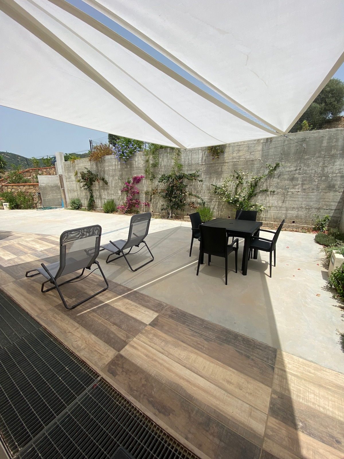 An outdoor patio area features a dining table with six black chairs, surrounded by a sunshade. Four reclining chairs are positioned nearby. The space is bordered by greenery and vibrant flowers, providing a serene environment for relaxation.