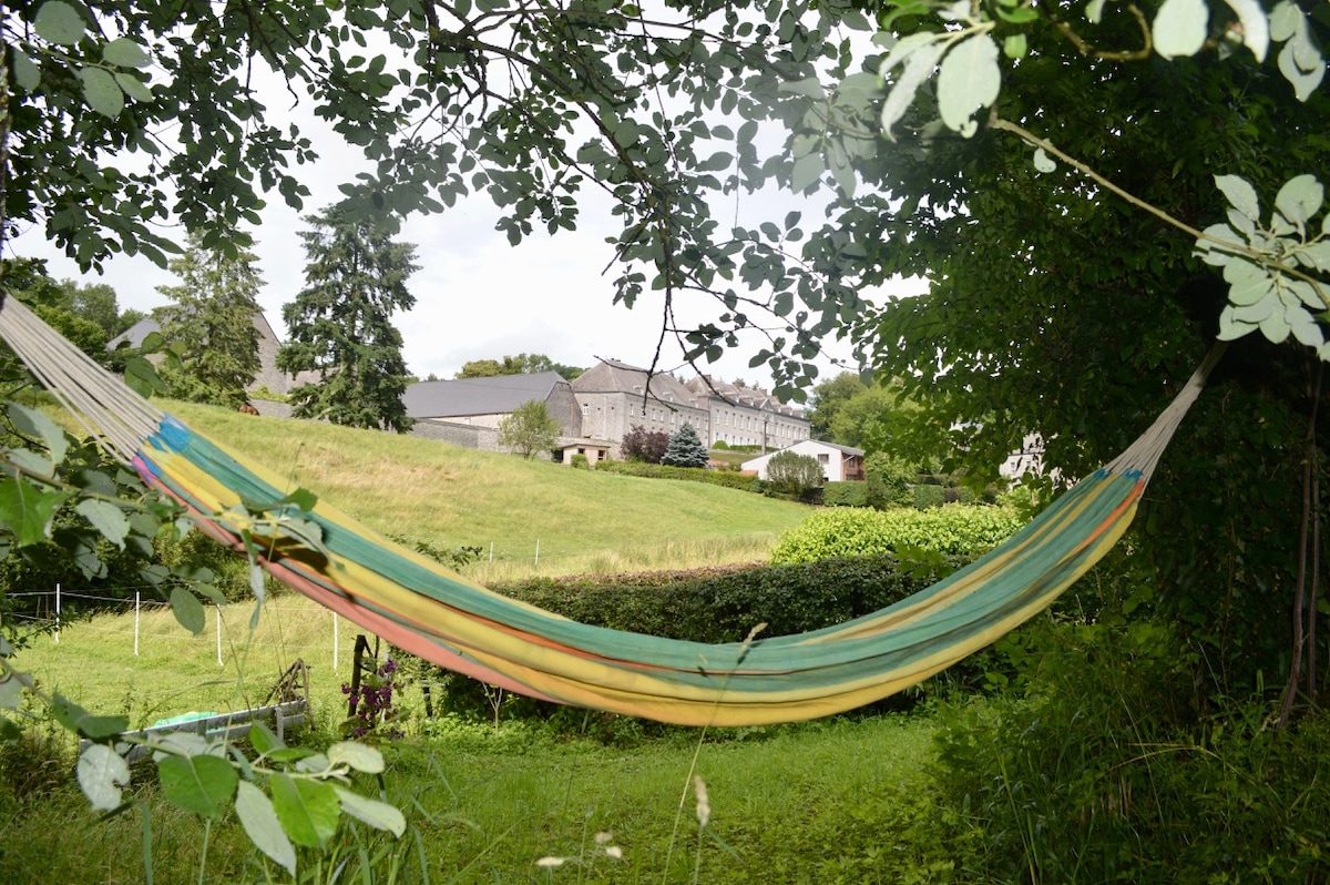 A colorful hammock is seen gently swaying between trees in a serene green landscape. In the background, a stone cottage is partially visible, surrounded by lush foliage and rolling hills. Natural light filters through the leaves, creating a peaceful atmosphere.