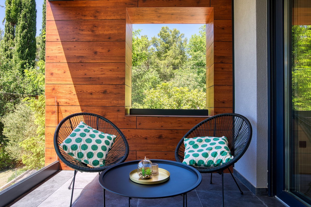 A cozy outdoor seating area features two black chairs with green and white patterned cushions, positioned around a round coffee table. A large, framed window offers a view of the lush greenery beyond, enhancing the serene atmosphere.