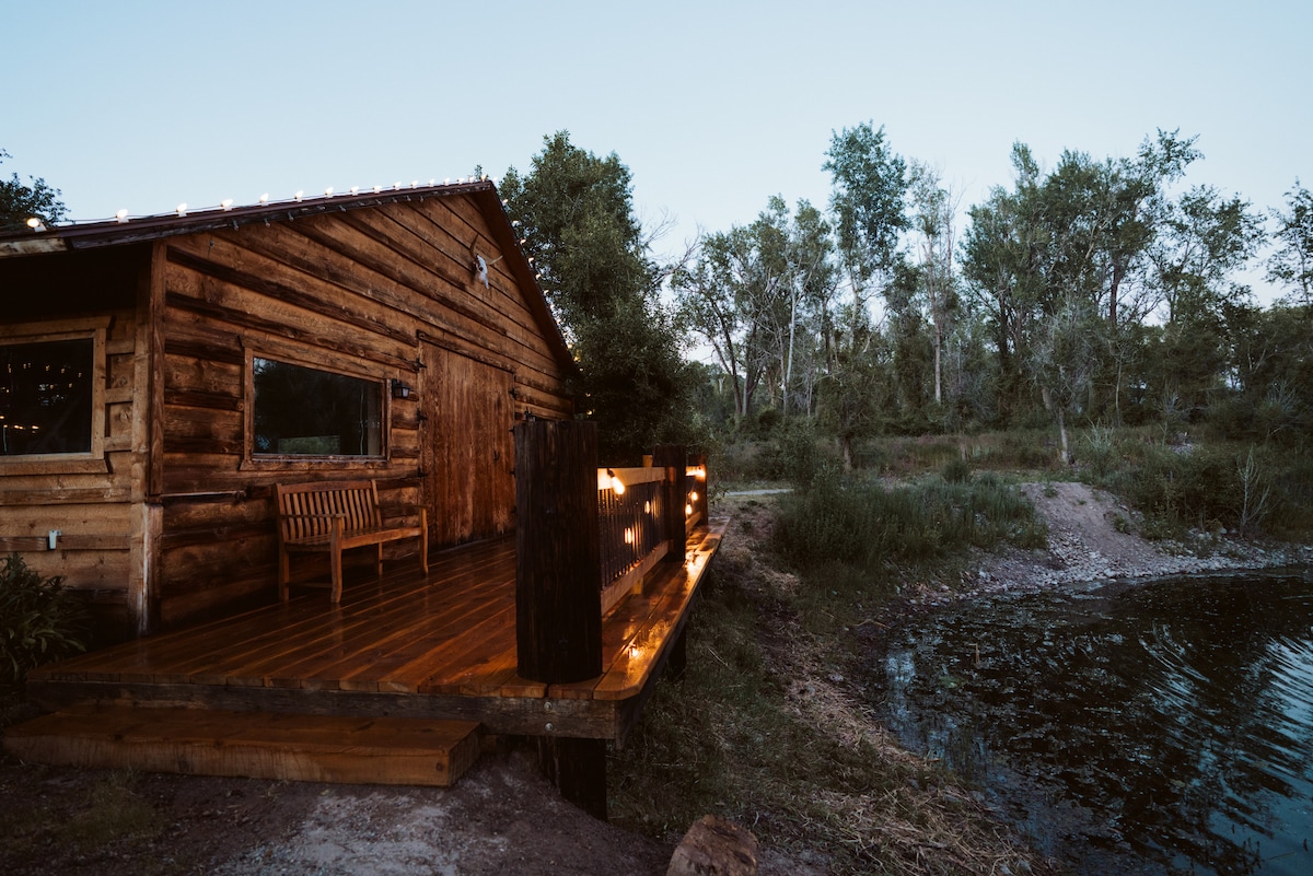 A rustic wooden cabin is positioned near a serene body of water, surrounded by greenery. A wooden bench sits on the porch, with a soft glow from string lights illuminating the entrance. Trees rise in the background, enhancing the natural setting.