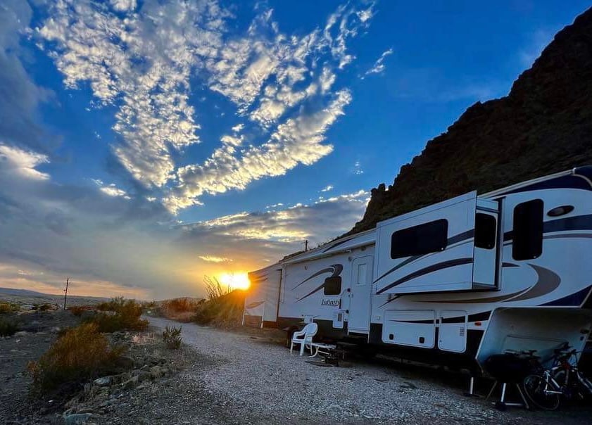 An RV is positioned amidst a natural landscape, with sunset light casting a warm glow on the exterior. The sky is filled with scattered clouds in shades of blue and orange, while gravel and desert plants surround the site.