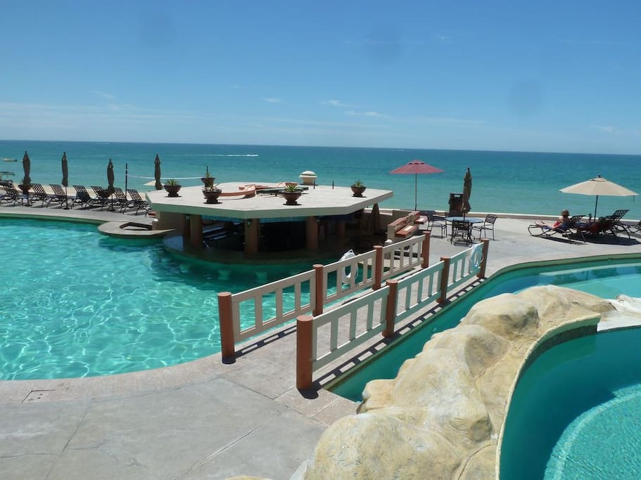 A serene pool area is highlighted, featuring a central swim-up bar surrounded by crystal-clear water. Lounge chairs line the pool's edge, with umbrellas providing shade. The ocean waves are visible in the background, blending seamlessly with the bright blue sky.