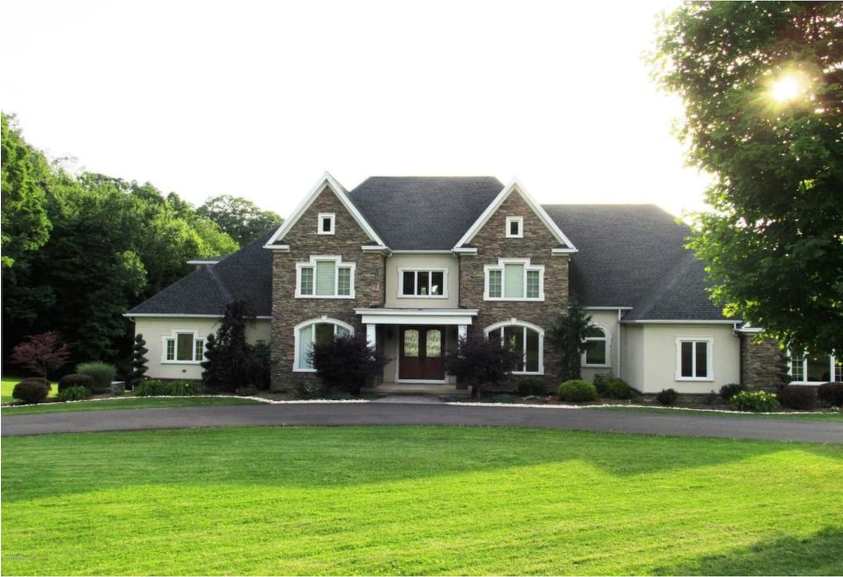 A large, elegant house is set on a manicured lawn, featuring a stone facade and a symmetrically designed entrance. Windows are adorned with white shutters, and lush greenery surrounds the property, creating an inviting exterior.