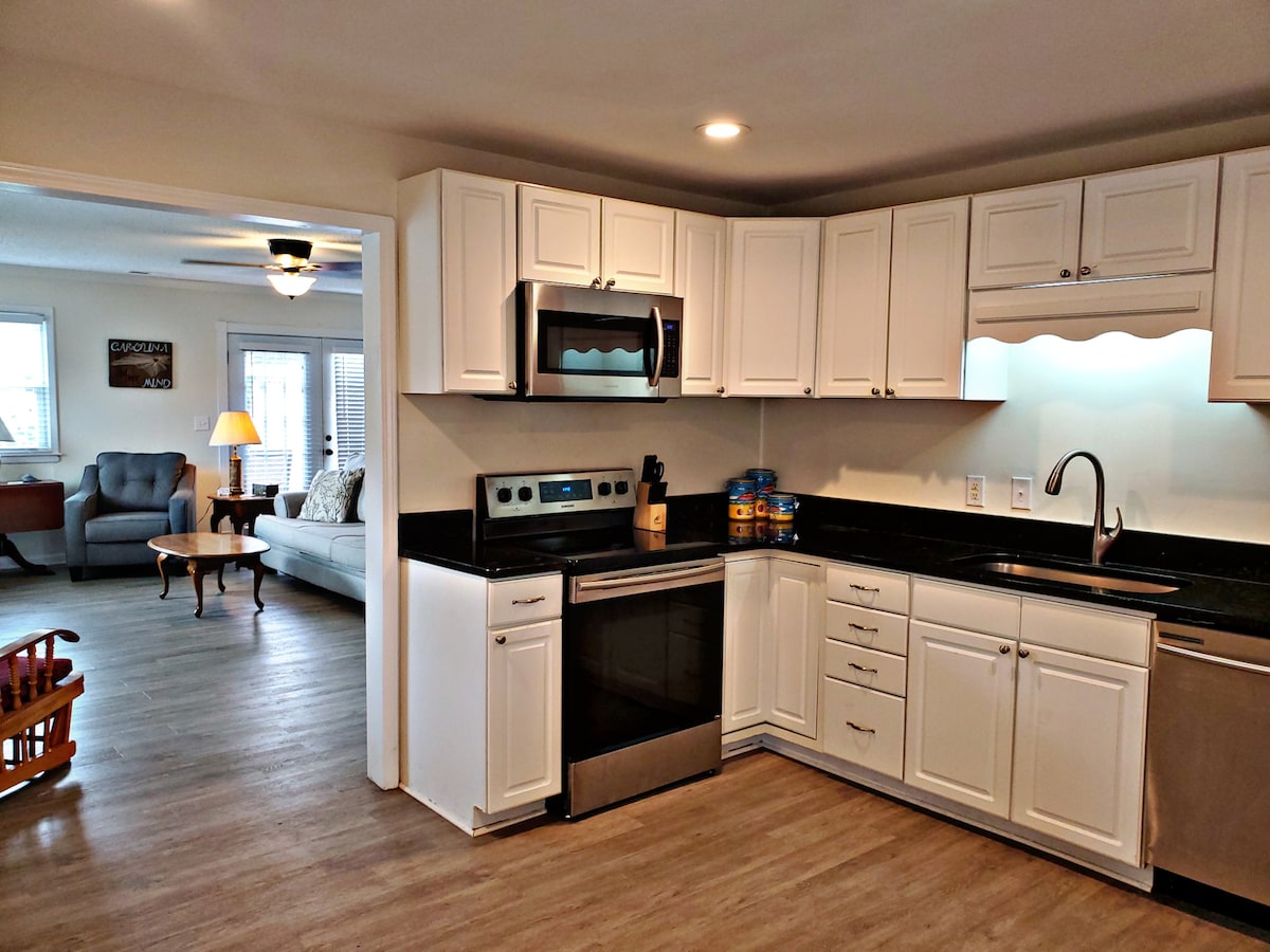 The kitchen features modern white cabinetry and sleek black countertops. Stainless steel appliances, including a microwave and dishwasher, are visible, along with a sink beneath under-cabinet lighting. The open plan reveals a cozy living area in the background, with comfortable seating arranged for relaxation.