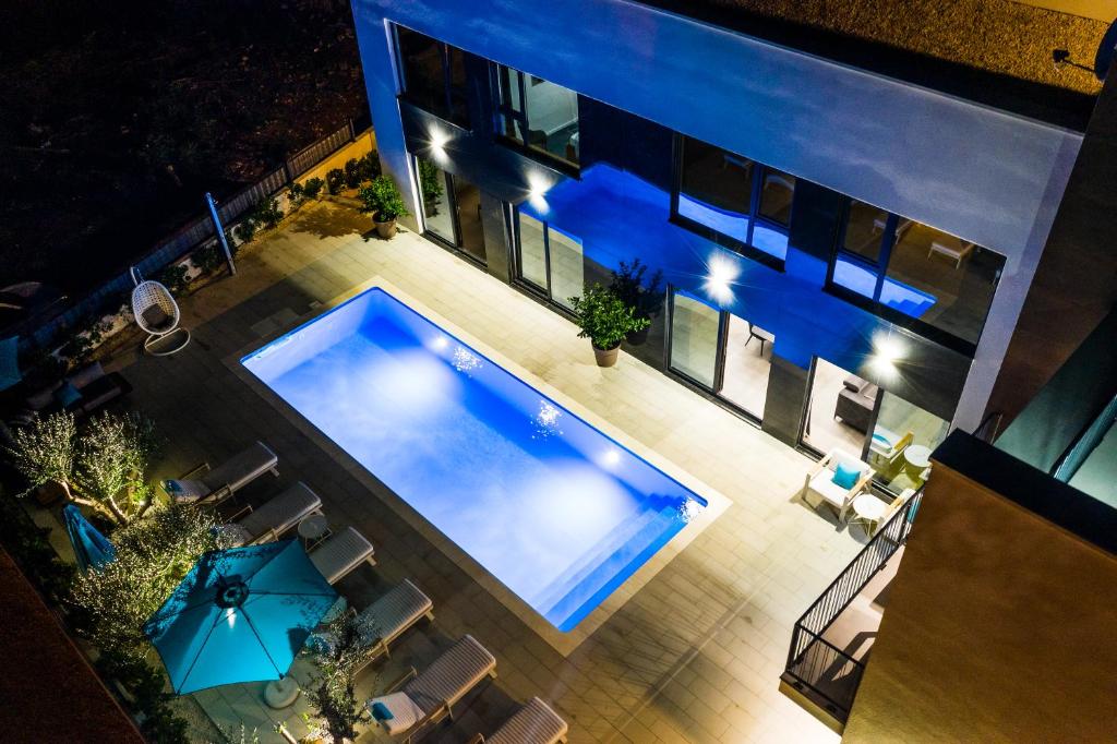 An aerial view of the outdoor area featuring a rectangular pool illuminated in blue light. Surrounding the pool are sun loungers, a large parasol, and potted greenery. Large glass doors provide a seamless connection to the interior, enhancing the open and airy feel.