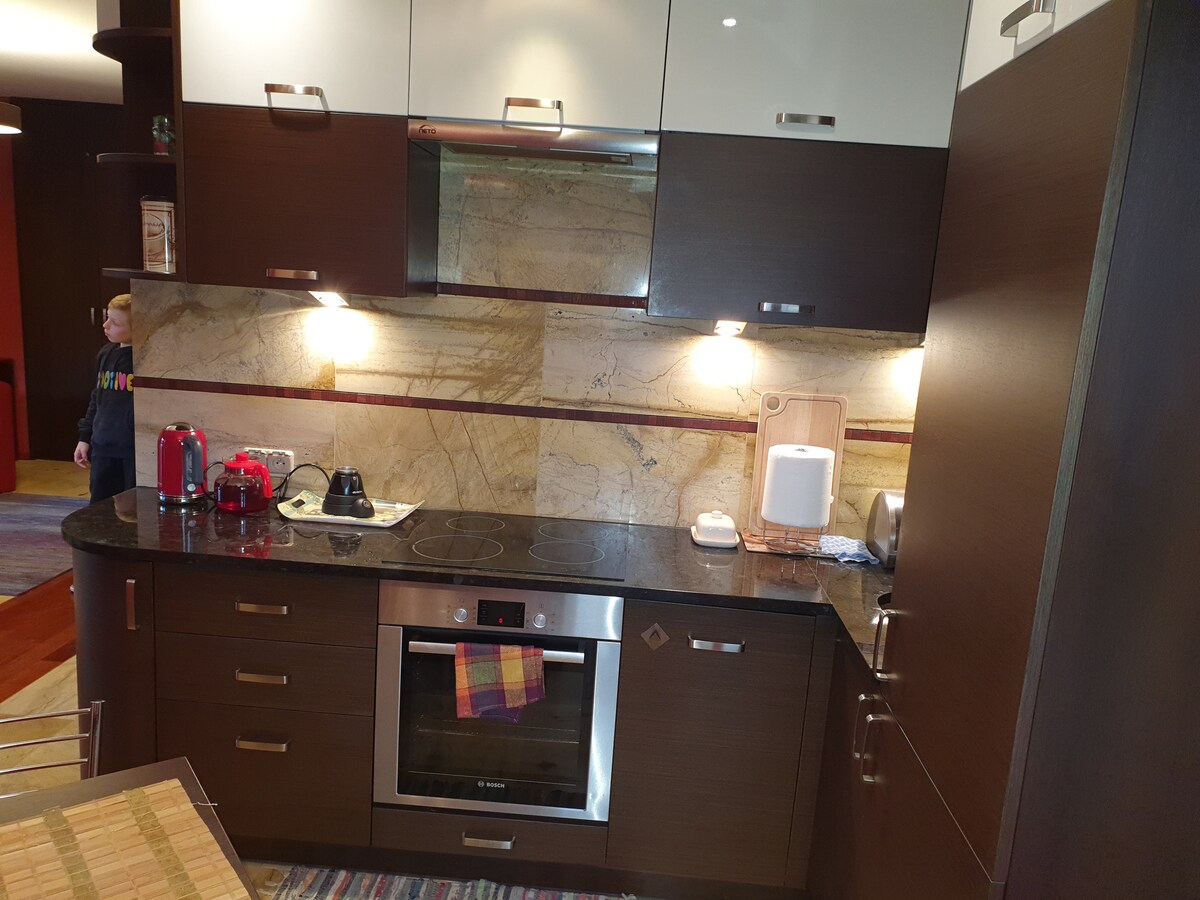 A modern kitchen is presented with dark wood cabinetry and light-colored upper cabinets. A granite countertop holds a kettle, toaster, and various appliances. Under-cabinet lighting enhances the workspace. The kitchen features an oven and a stovetop, integrating functionality with a sleek design.